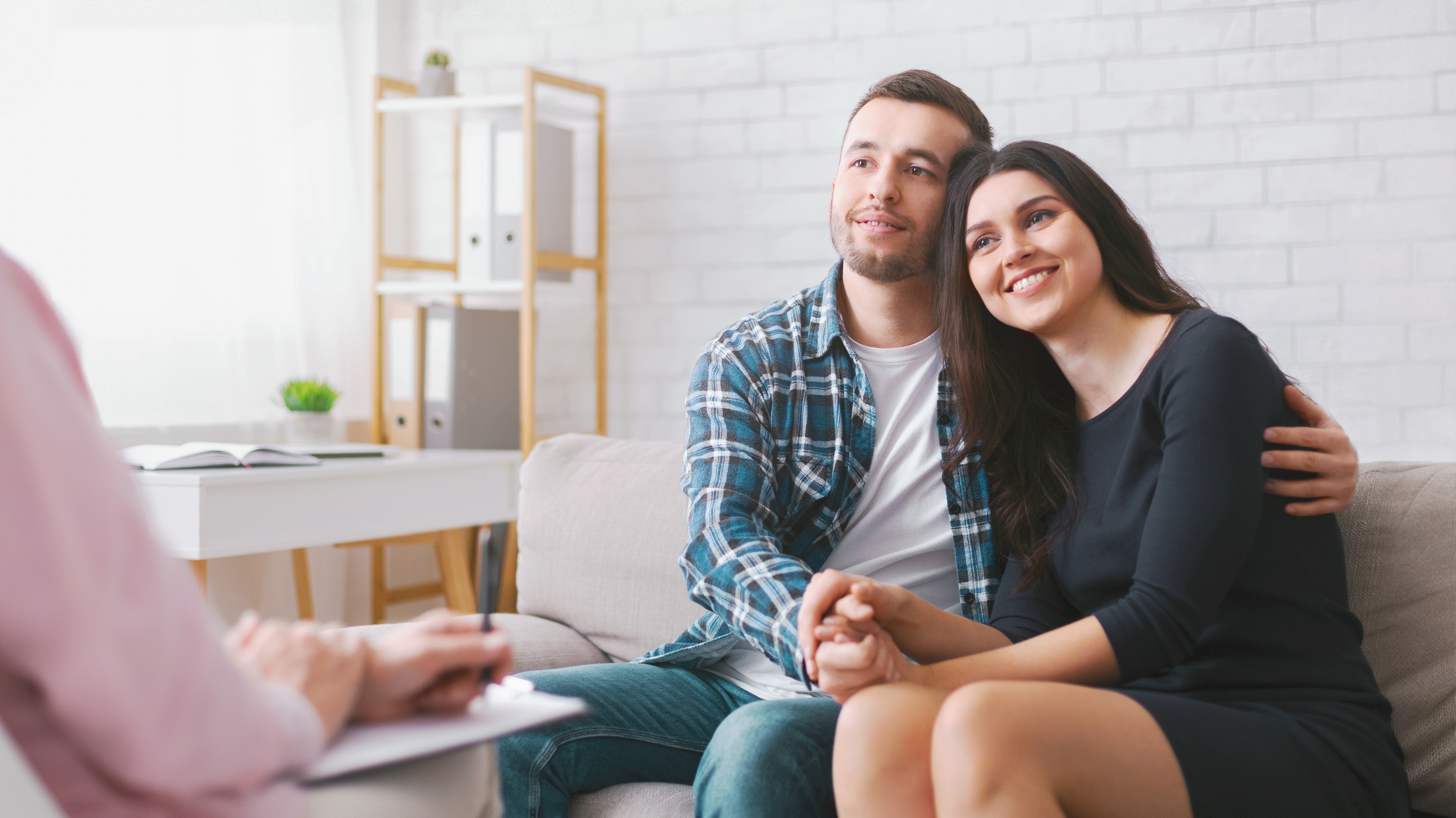 Ein Paar sitzt auf einer Couch während einer Therapiesitzung, hält sich an den Händen und blickt nachdenklich zur Seite.